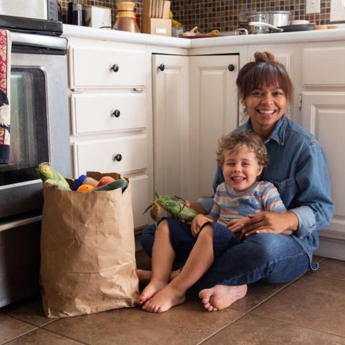 A woman and child sitting in a kitchen with a bag of groceries