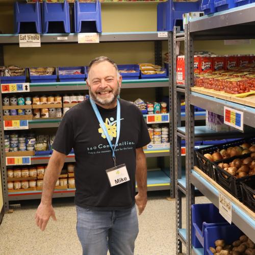 Volunteer Mike in 360 Communities Food Shelf Smiling volunteer with beard in a 360 Communities shirt surrounded by shelves of food.