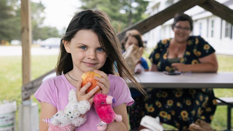 A child holds an orange while looking at the camera. Two people in the background are seated at a picnic table.