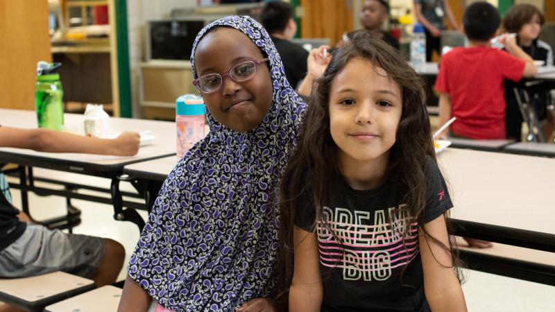 Two kids smile while sitting at lunchroom table
