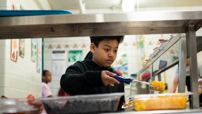 A young boy scoops a serving of oranges onto his school lunch tray