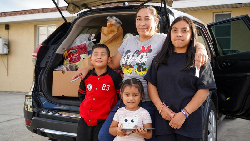 Mom with arms around 3 kids, standing in front of car filled with food