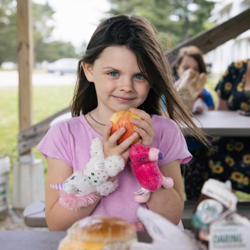 A child holds an orange while looking at the camera. Two people in the background are seated at a picnic table.