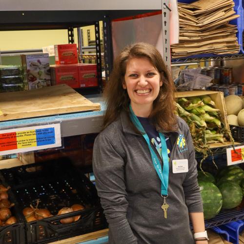 A brunet woman in gray smiling at the camera in front of shelves of food