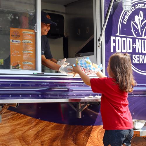 A child in a red shirt accepts a meal from a person through a food truck window