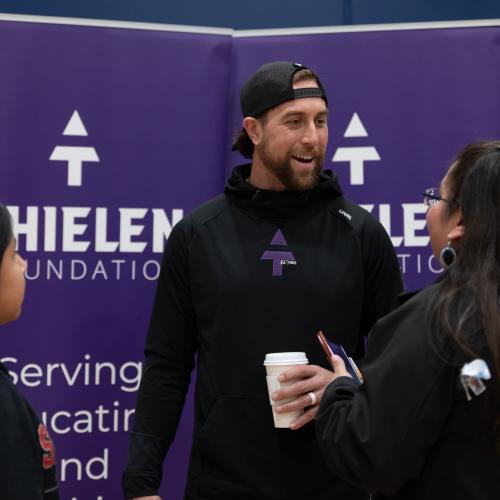 A man wearing a black shirt and hat smiling and talking in front of purple Thielen Foundation signs