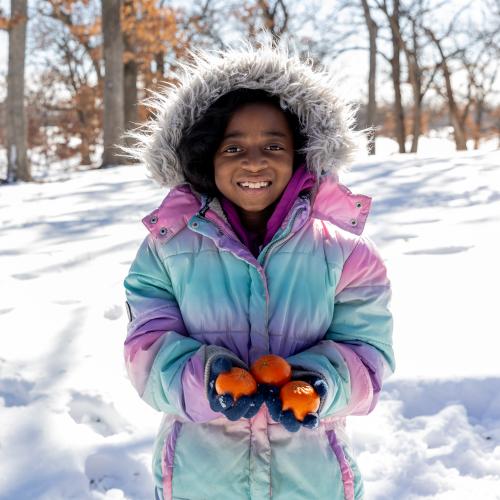 Young girl outside in the snow in her winter coat holding oranges