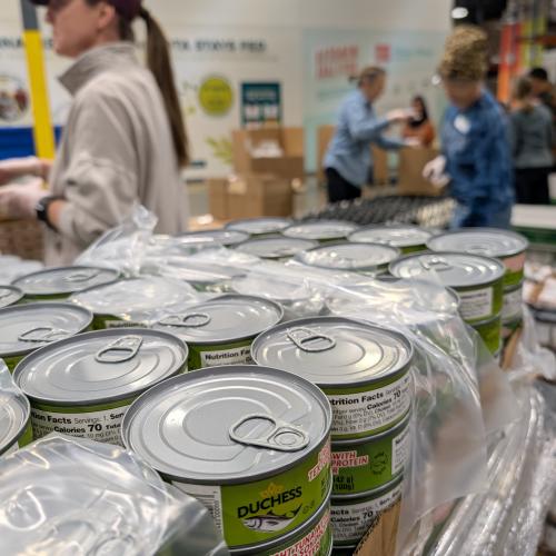 Stacks of tuna cans in the foreground and volunteers packing boxes in the background