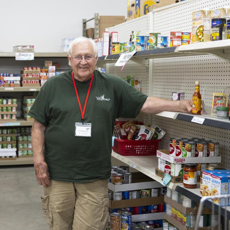 A man smiles at the camera and stands in front of shelfs of canned and boxed food