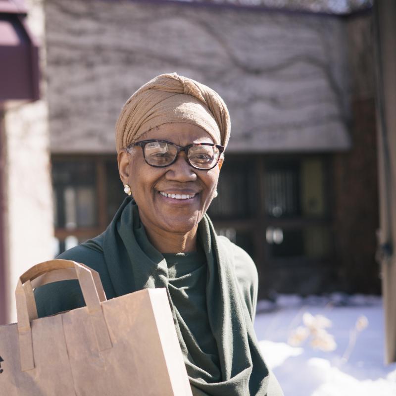A senior woman with glasses carries a brown paper grocery bag