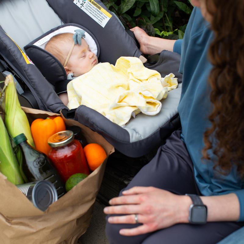 A baby in a car seat next to a bag of groceries with a mother looking down on the child.