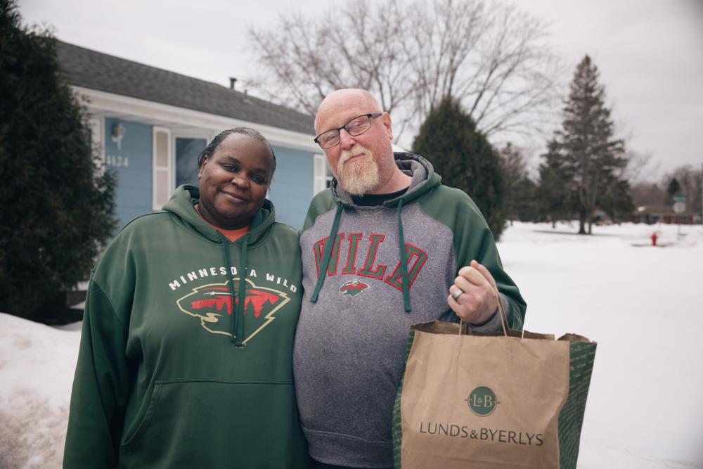 Two people standing outside, smiling and holding a grocery bag