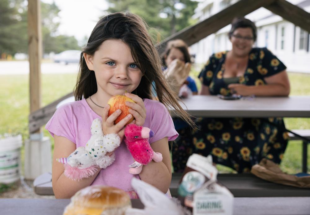 A child holds an orange while looking at the camera. Two people in the background are seated at a picnic table.