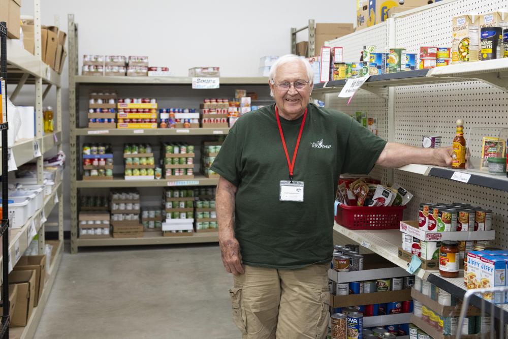 A man smiles at the camera and stands in front of shelfs of canned and boxed food