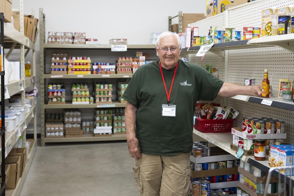 Buffalo Food Shelf A man smiles at the camera and stands in front of shelfs of canned and boxed food