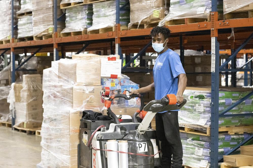 An employee rides a pallet jack filled with boxes while smiling at the camera