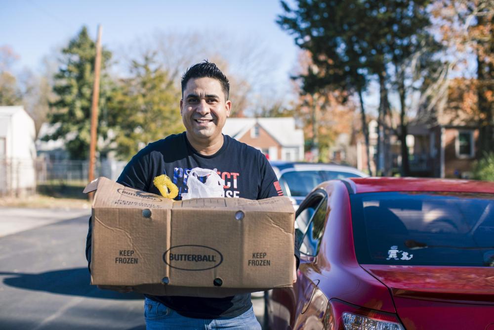 A person holds a box while standing next to a car and smiling at the camera