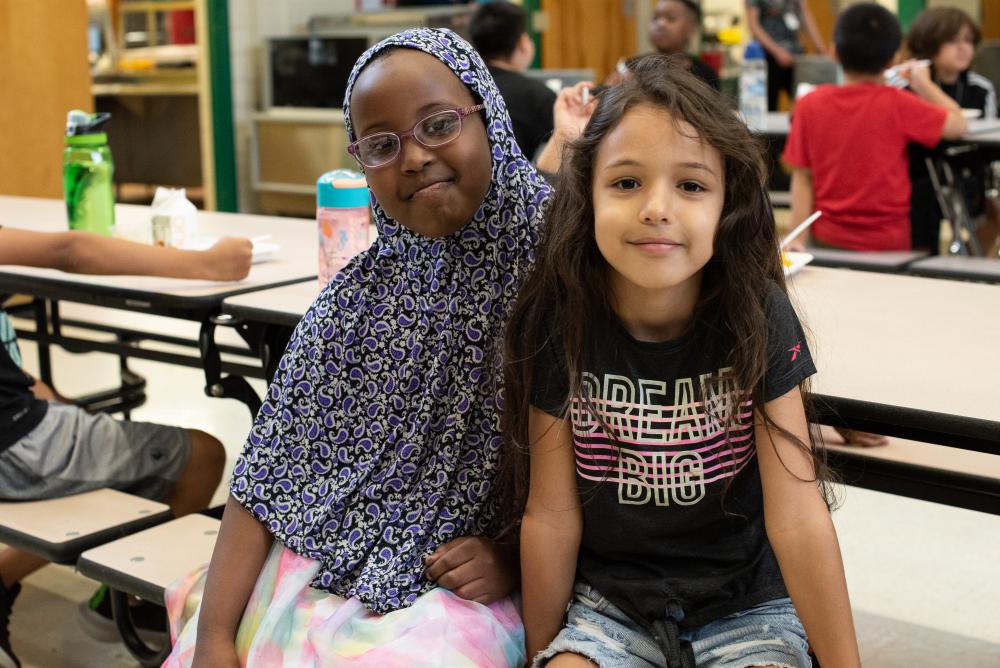 Two kids smile while sitting at lunchroom table