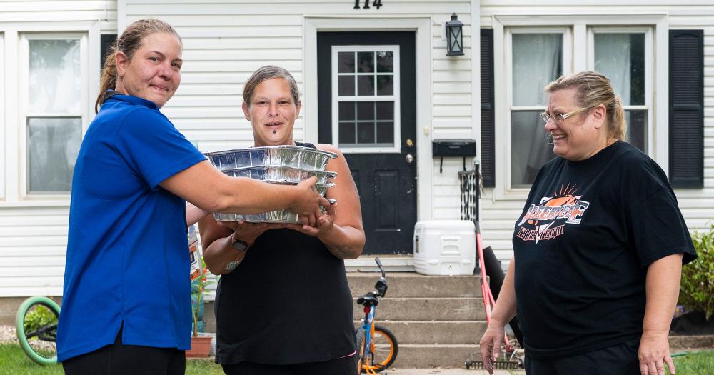 three women standing outside their home holding containers of prepared food