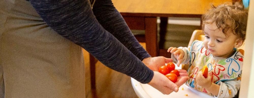 Support families with monthly giving Toddler being handed tomatoes in a high chair