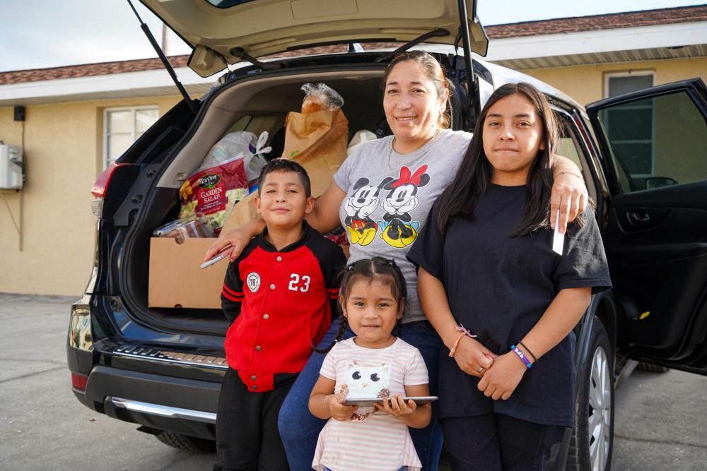 Mom with arms around 3 kids, standing in front of car filled with food
