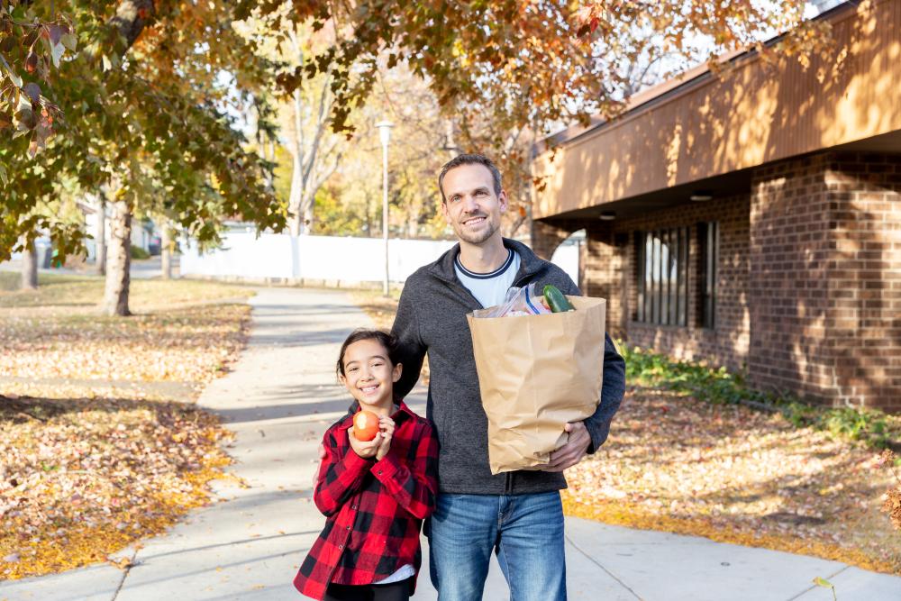 24hour Harvest Child and adult standing next to each other. Child has apple in hand and adult is holding grocery bags