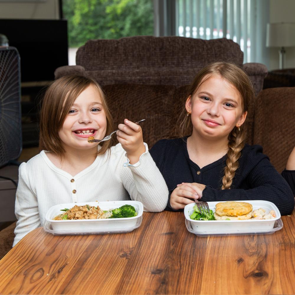 Two children sitting at a table inside, eating prepared meals