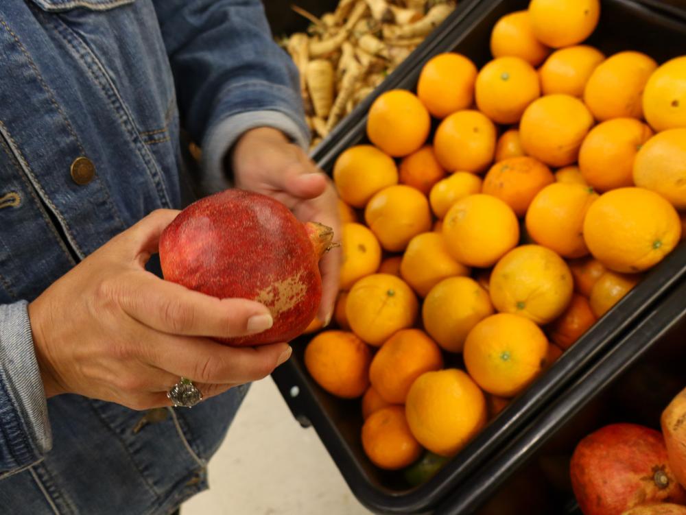 Rhory holding produce at ACBC Food Shelf A woman's hands holding a pomegranate in front of a rack of oranges at ACBC Food Shelf