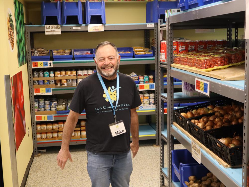 Volunteer Mike in 360 Communities Food Shelf Smiling volunteer with beard in a 360 Communities shirt surrounded by shelves of food.