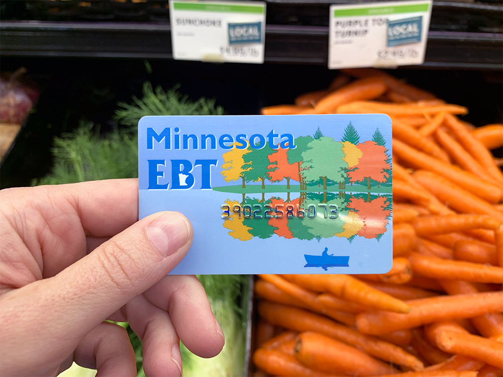 A white hand holding a Minnesota EBT card in a grocery store produce section