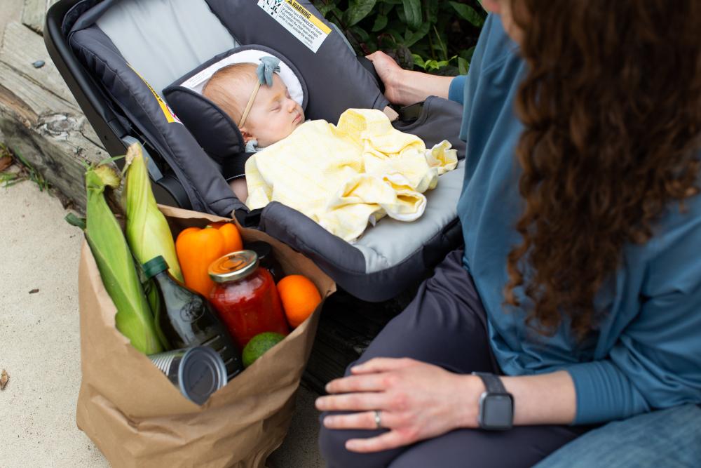 A baby in a car seat next to a bag of groceries with a mother looking down on the child.