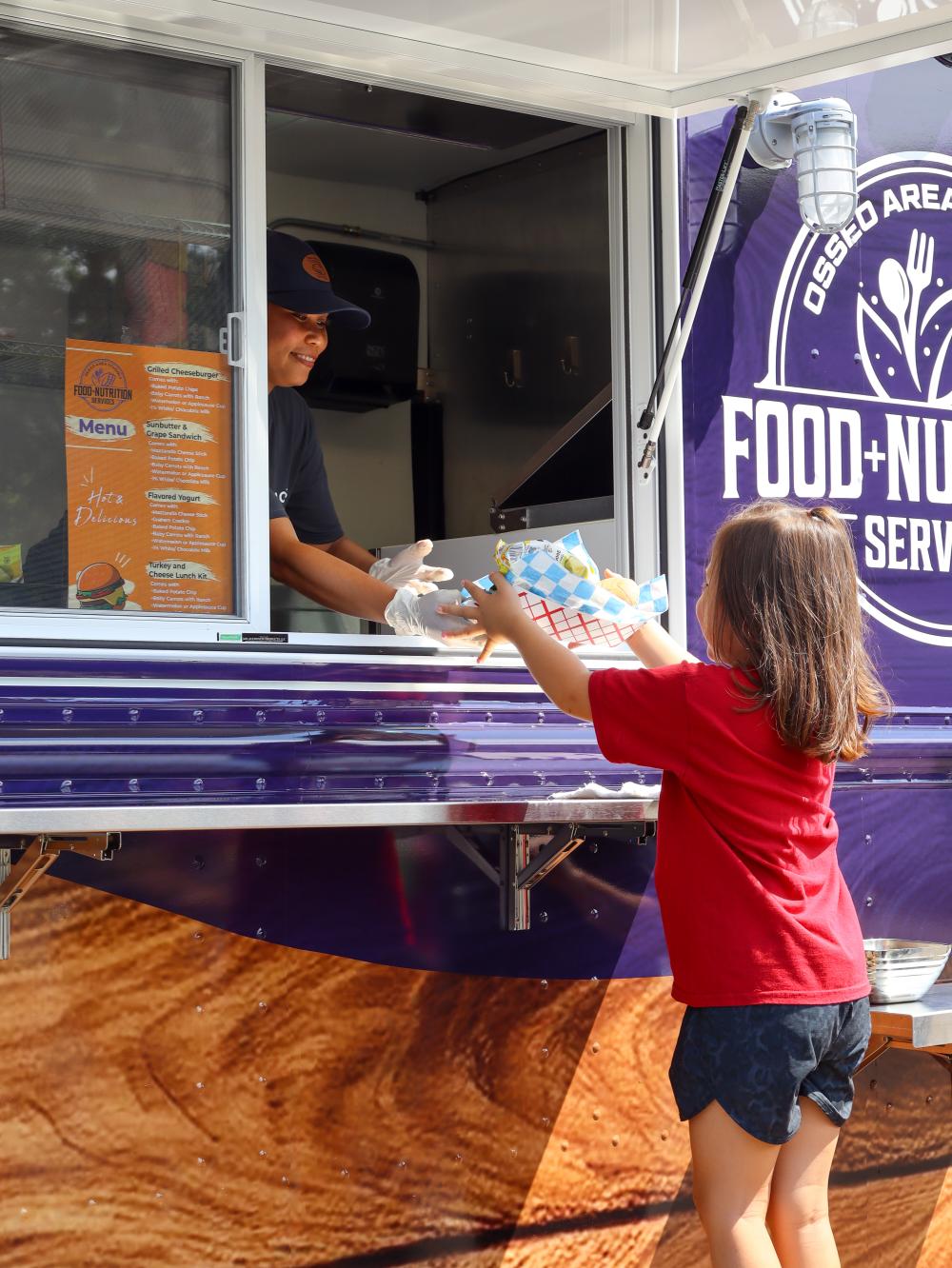 Osseo Area Schools Food Truck A child in a red shirt accepts a meal from a person through a food truck window