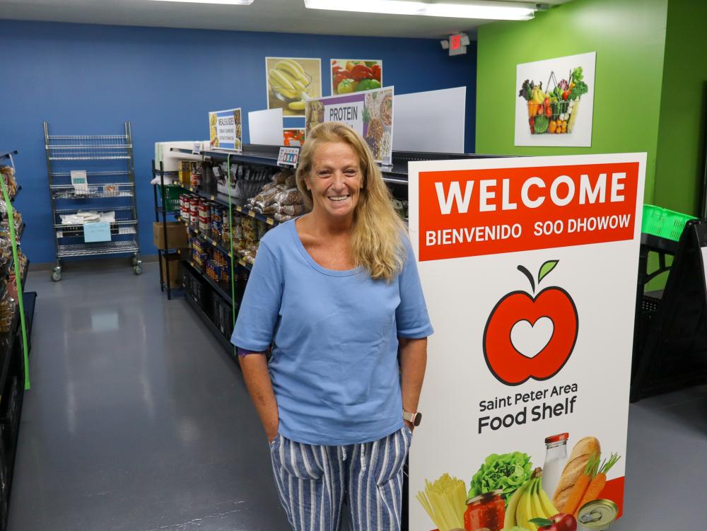 Jodi in St. Peter Area Food Shelf woman in blue shirt standing beside a welcome sign next shelves of food in food shelf