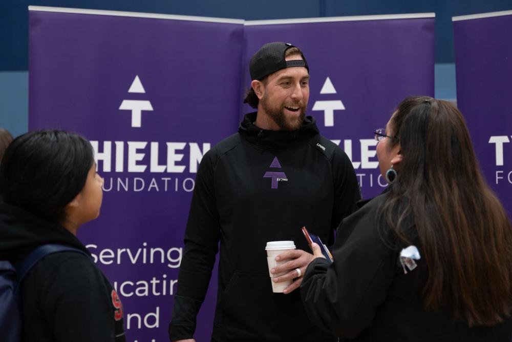 A man wearing a black shirt and hat smiling and talking in front of purple Thielen Foundation signs