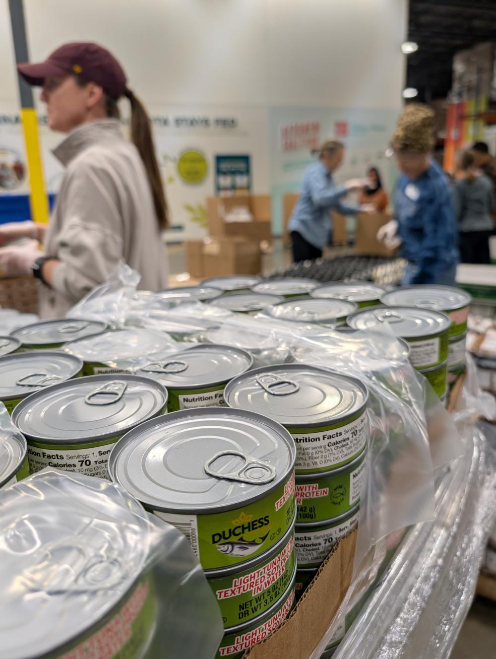 Stacks of tuna cans in the foreground and volunteers packing boxes in the background