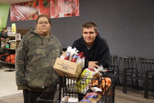 A mother and son pose with a shopping cart filled with bread and produce.