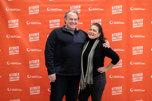 A man and a woman in black shirts pose in front of a red Kitchen Coalition mural