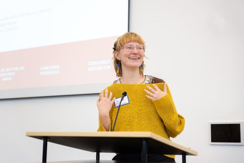 A woman in a yellow sweater speaks at a podium