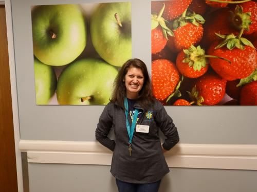 Sara Felix A brunet woman with a gray zip up and lanyard posing in front of photos of apples and strawberries.