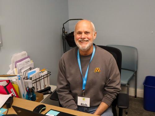 Volunteer Rich at 360 Communities Volunteer with a white beard in gray UMN sweatshirt sitting at a desk
