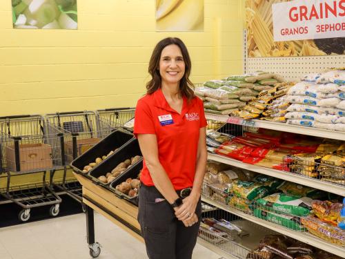 ED Stephanie Lehman, a brunette woman in a red shirt, stands in front of produce in the food shelf, smiling at the camera