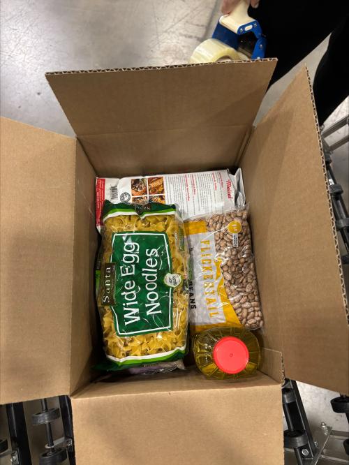 An open cardboard box containing egg noodles, beans, oil, and other groceries