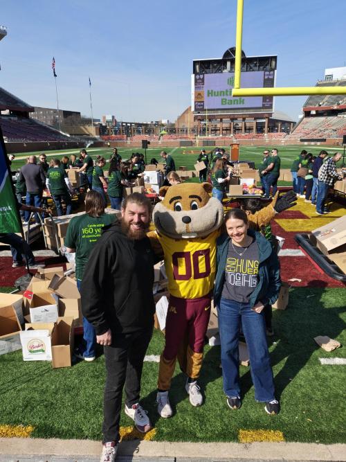 Two people pose with UMN Goldy the Gopher mascot in front of volunteers working.